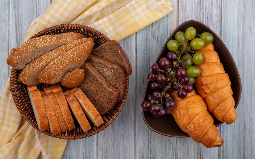 top-view-breads-as-sliced-seeded-brown-cob-rye-crusty-ones-basket-plaid-cloth-bowl-croissants-grapes-wooden-background_141793-27721